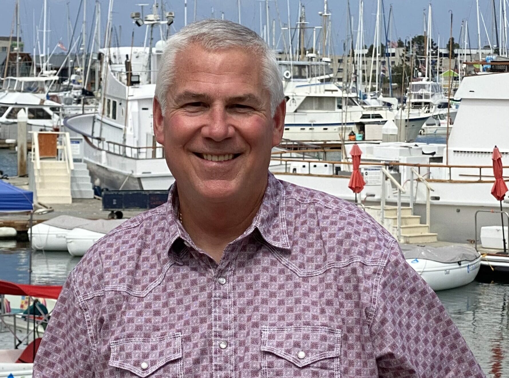 Cary Attl in a purple western style shirt leans against white railing in front of yachts.