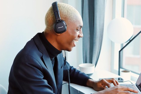 A young man, wearing headphones, while smiling as he watches something on his laptop.