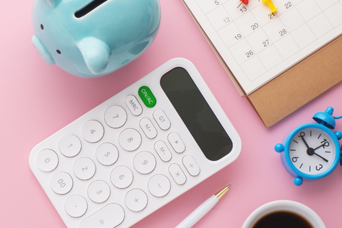 A tidy arrangement of a piggy bank, calculator, calendar, alarm, coffee and pen amid a soft pink background