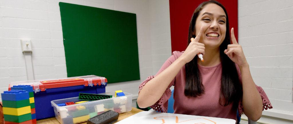 A young adult Latina who is a speech language pathologist shows all of her teeth as she holds two fingers upward. She is surrounded by toys.