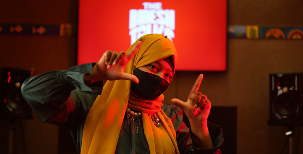 Anika wears a bright yellow hijab and black face mask in front of a glowing red display of The Bidesh Talks Logo.