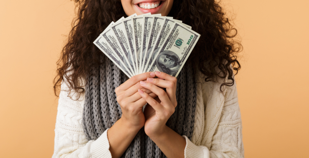 A woman with dark curly hair and mocha brown skin holds up a row of hundred dollar bills
