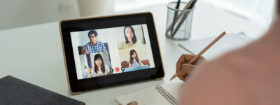 A person takes notes while watching four people in a virtual meeting on a tablet screen.