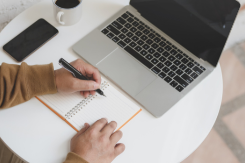 Aerial view of a round white table, covered with an open laptop computer with a blank screen. To the left of the laptop is a cup a coffee and a smartphone. In front of the computer is a person with only their hands visible, writing in an orange notebook with their left hand.
