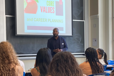 Akunna Rosser presents to the class. Behind Akunna is a projector screen reading,