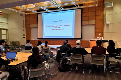 A certificate program presenter stands at the podium in the front of a lecture hall, speaking to a group of engaged attendees.