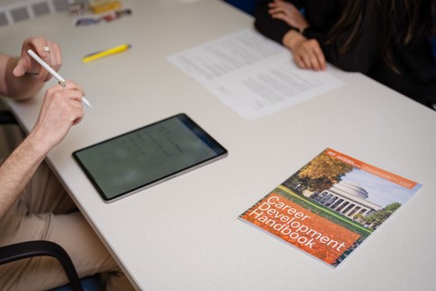 Two people working across from each other at a table with papers, a tablet, and a career development handbook; only their hands are visible.