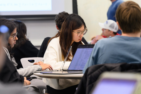 Students work on laptops at round tables during an MIT class focused on computer-based learning, with a projected screen visible at the front of the room.