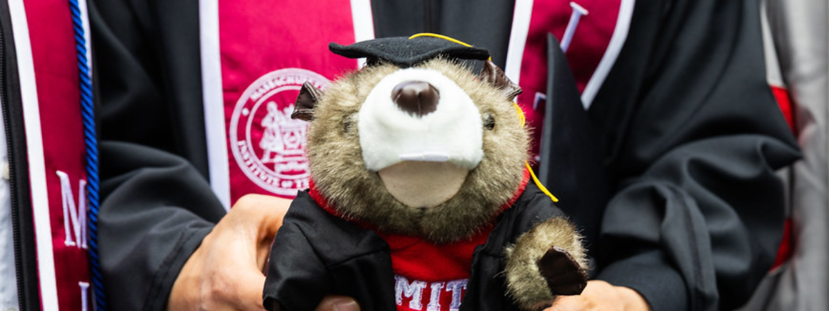 A graduate holds a Tim the Beaver plush wearing full graduation regalia.