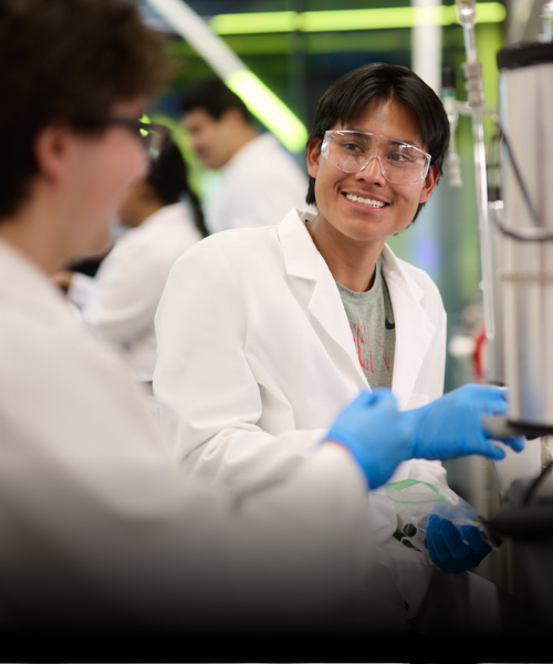 Student in a lab coat smiling at another student in a university lab.