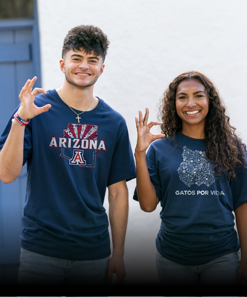 Two students in blue t-shirts with University of Arizona branding smile at the camera and hold up the Wildcat hand gesture.