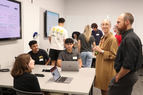 Four students sit at a table behind their laptops and present a slideshow to two adults standing by.