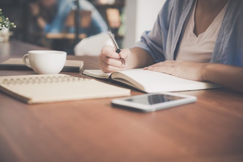 Person holding a pen over a blank notebook. The notebook sits on a table next to a phone, another notebook, and mug
