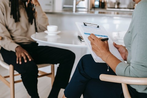 Two people shown sitting at a table but you are unable to see their faces. One is holding a clipboard.