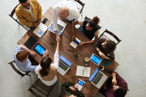 Aerial view of eight people sitting around a table with laptops and notebooks. Two of the eight are shaking hands across the table.