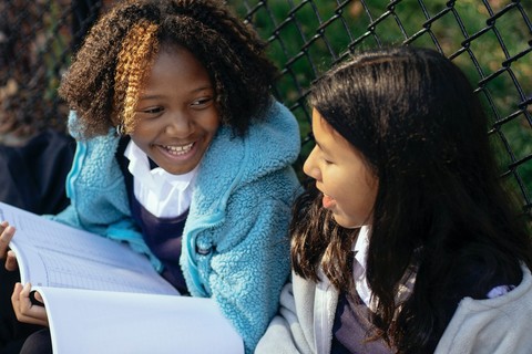 Two preteens sit outside with workbooks on their laps talking
