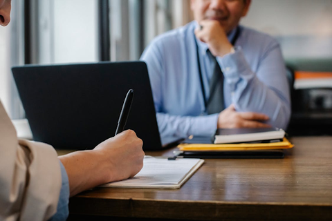 Photo of people sitting across the table from one another. One person is writing while the other rests their chin in their hand, sitting behind an open laptop.