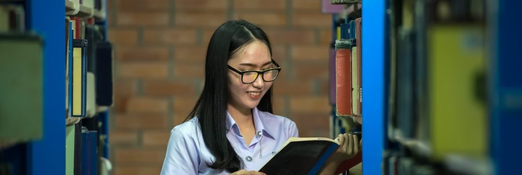 woman with glasses reading a book between two book cases in a library