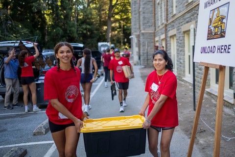 Move-in and orientation day for first year students on the campus of Swarthore College. Two students are shown carrying a box in front of Wharton Hall. In the foreground is a sign reading
