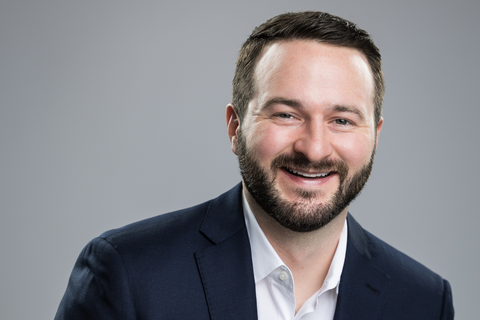 Ryan has short, dark hair and a beard. He wears a professional blazer and button-down while smiling at the camera, against a gray background.