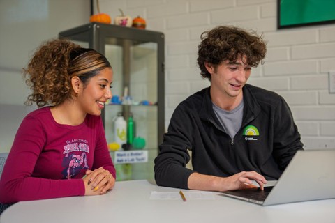 A student leader and a peer look at a computer at a work table together.