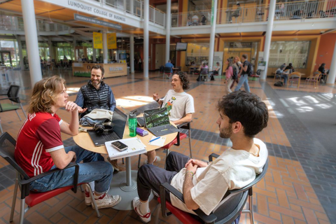 Four students socialize at a round table in the Lillis atrium. They have notebooks and laptops out, and are deeply engaged in conversation.
