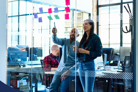 People speaking and pointing at post-its on a clear glass wall in a boardroom.