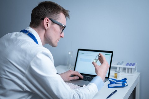 A young medical professional is staring at a vial while entering information into a computer system.