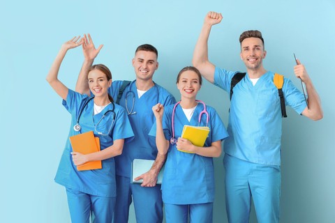 Group of nurses wear scrubs standing together while holding books and smiling in front of a blue background.