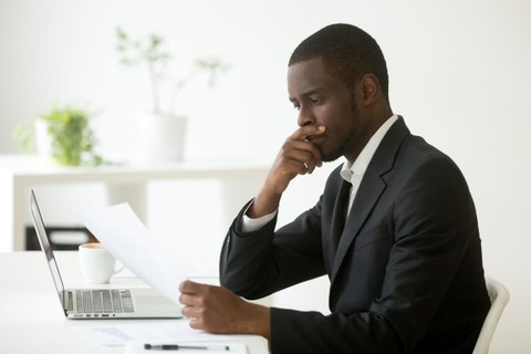 Serious african-american businessman employer thinking of business offer reading mail cover letter at workplace, puzzled black company executive looking at financial document considering contract.