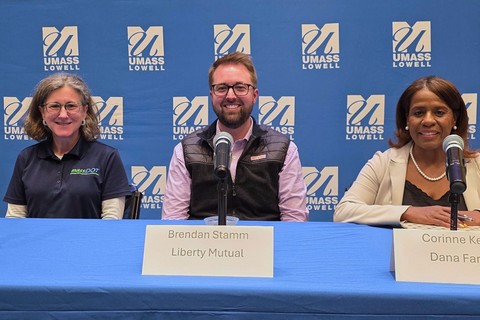 Three panelists sit behind microphones at a UMass Lowell event, with name placards for Brendan Stamm and Corinne Kelton.