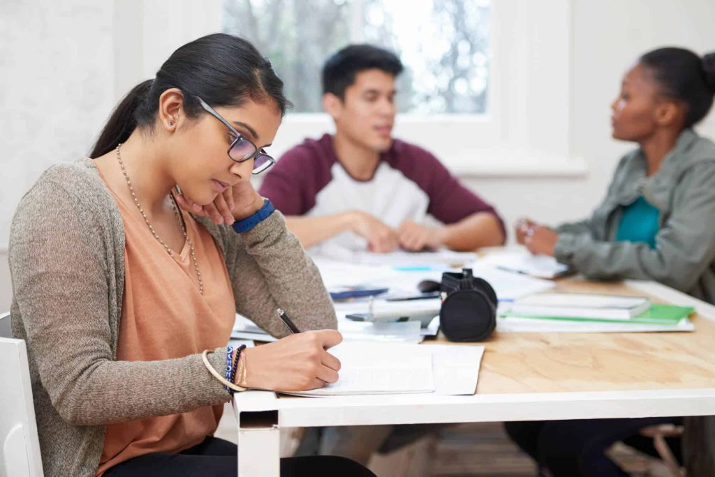 Three college students sit around a table. The student in the foreground writes in a notebook. The two students behind her are in the middle of a conversation. All three students look engaged and interested in what they're doing.