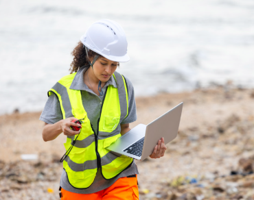 Person wearing safety gear holding a computer on a beach with trash on it.
