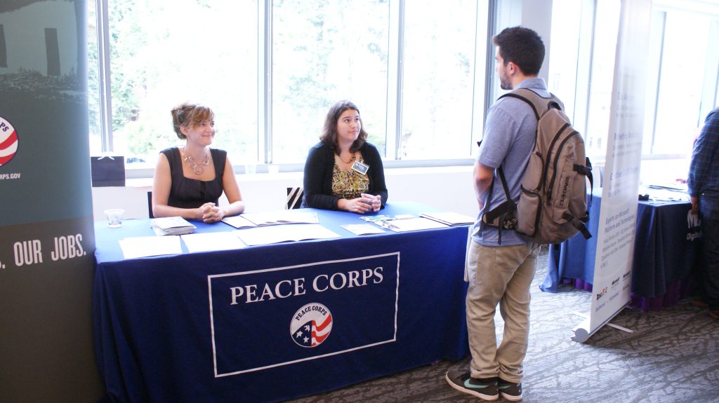 Two employers are meeting with a student at a career fair. The employers are both female-presenting and are seated at their table, covered with a drape that says "Peace Corps." The student is standing and is male-preseting, wearing a blue short-sleeve dress shirt and khaki pants.
