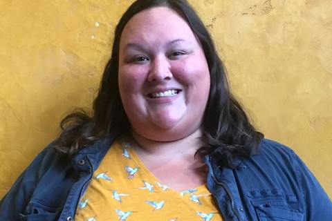 A native woman with brunette hair in a yellow floral shirt and a blue utility jacket standing in front of a yellow background.