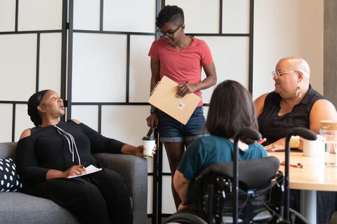 4 people sitting around a table chatting. One person with brown hair in a wheelchair, one bald woman sitting, one Black woman sitting looking up at another Black woman standing with a folder in her hand.