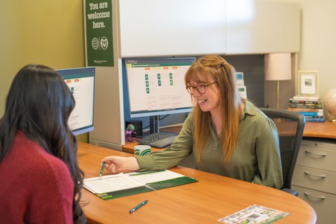 A career counselor is seated at a desk, smiling and holding a pen while pointing to documents during a conversation. Two computer monitors are visible behind them, and a sign on the wall reads 'You are welcome here.' The office setting includes books, a lamp, and other desk items.