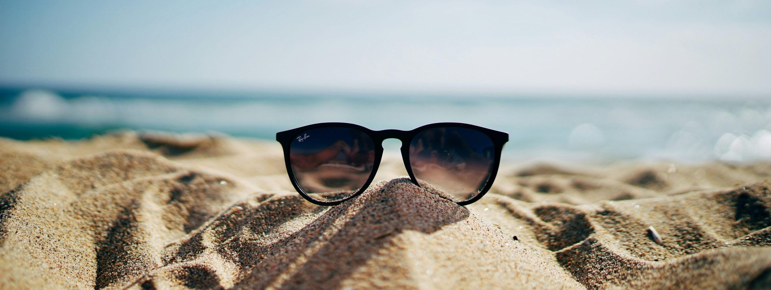 Sunglasses propped on a pile of beach sand with the ocean in the background.