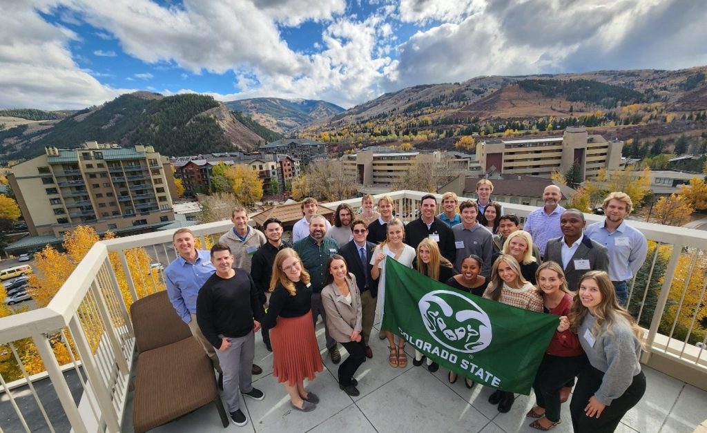 Students in business causal attire holding CSU flag stand on a hotel patio overlooking mountains covered in yellow aspen trees and green pines. 