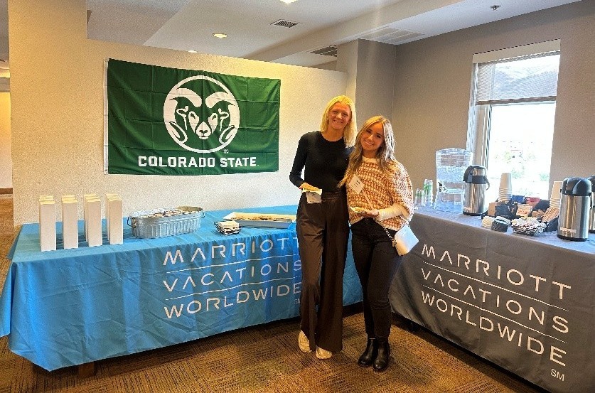 Two CSU students stand holding refreshments in front of two tables with coffee, cookies, and gifts from Marriott Vacations Worldwide.