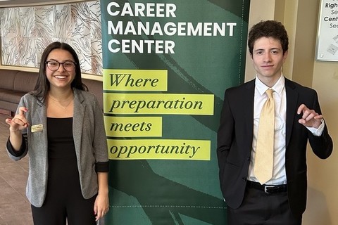 Two Student Career Navigators standing beside a banner that reads 'CAREER MANAGEMENT CENTER' and 'Where preparation meets opportunity.' One wears glasses, a gray blazer, and black pants; the other wears a black suit with a white shirt and yellow tie. Both are making expressive hand gestures mimicking ram horns.