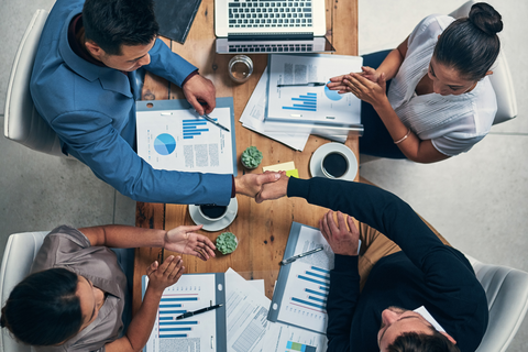 Overhead view of four people seated around a wooden table with documents, charts, and a laptop, two shaking hands.
