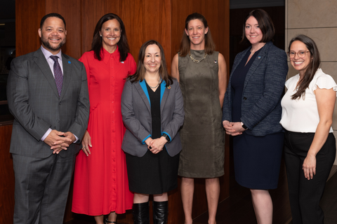A group of six people in business attire stand indoors against a wooden backdrop, smiling at the camera.