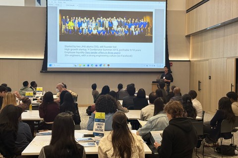 A presenter speaks to a large group of students seated at tables during a career event, with a presentation about a startup company projected on a large screen.