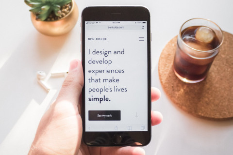 Image of a hand holding a phone over a desk with a plant and drink in the background with the text: I design and develop experiences that make peoples live simple.