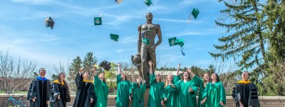 Spartan graduates standing in front of Sparty statue on campus throwing graduation caps in the air