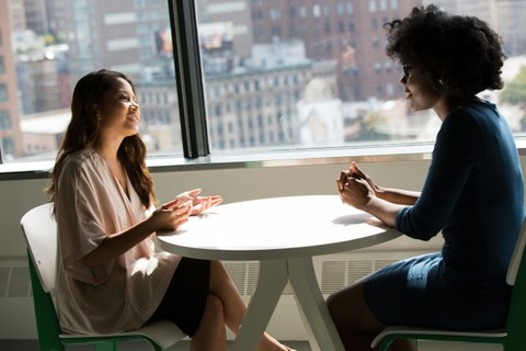 Two women talking at a table in front of a high-rise window.