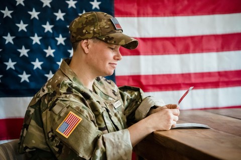 Army serviceperson sitting at a desk in front of a US flag, looking at a phone.