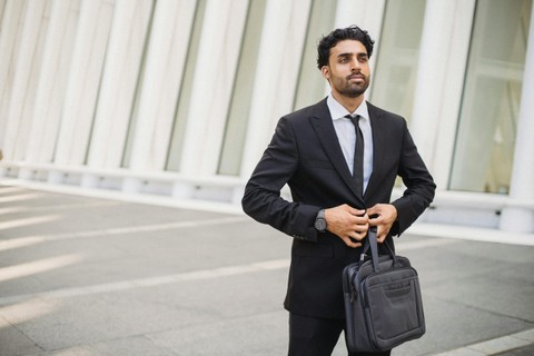 Man in suit, walking in front of white building and carrying a black computer bag.