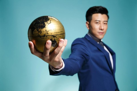 Man in blue suit holding a gold globe towards the camera; teal background